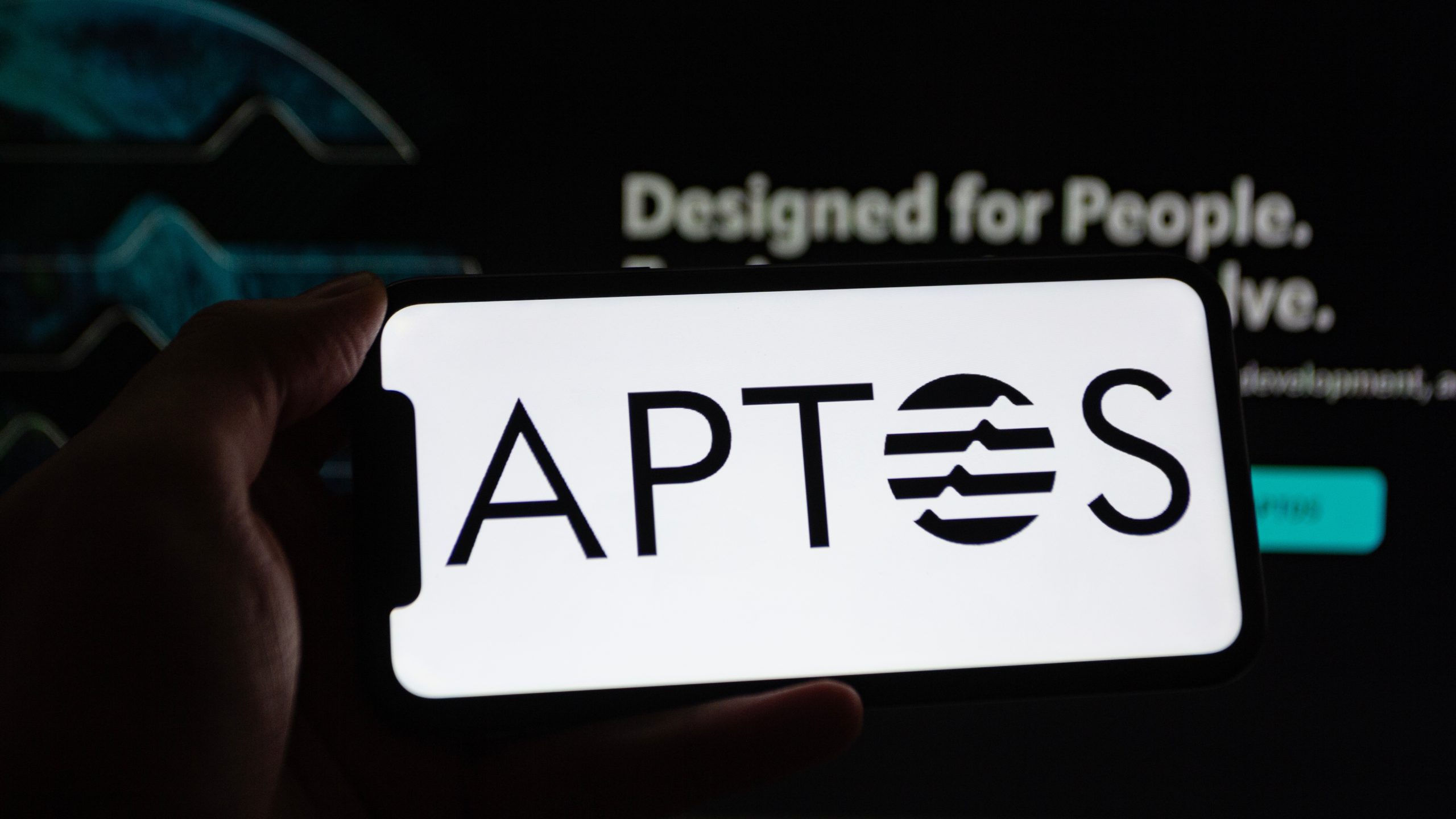 Ein Smartphone, das das Aptos-Logo in dunkler Umgebung mit unscharfem Text im Hintergrund anzeigt.