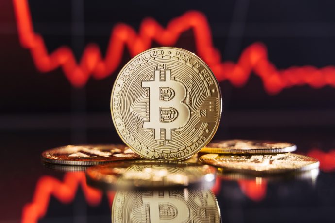Im Vordergrund ist ein Stapel Bitcoin-Münzen zu sehen, vor einem unscharfen Hintergrund mit einem nach unten geneigten roten Finanzdiagramm, das die Volatilität des Kryptomarkts widerspiegelt.