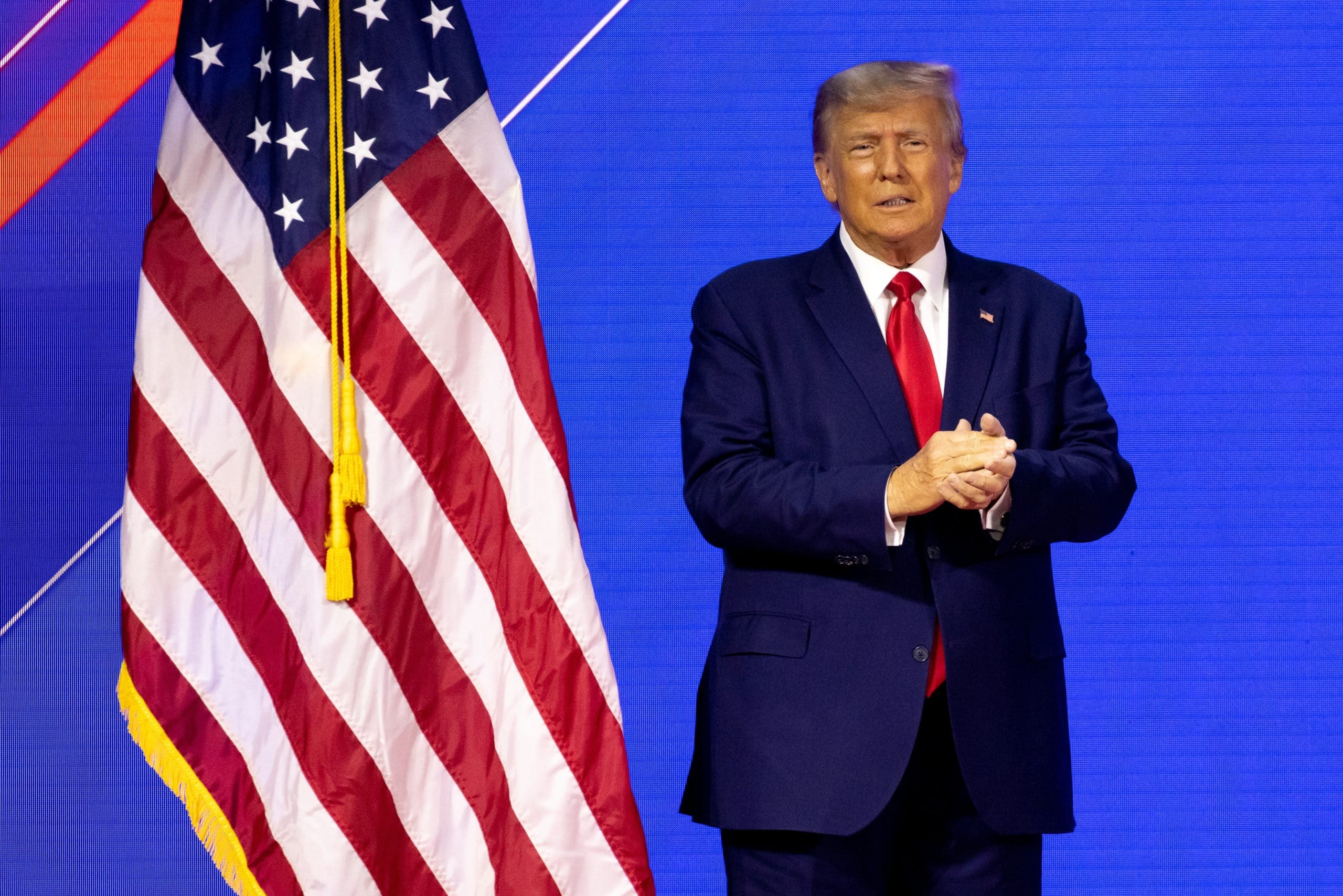 Donald Trump in einem marineblauen Anzug und einer roten Krawatte steht auf der Bühne neben einer US-Flagge vor einem blauen Hintergrund.