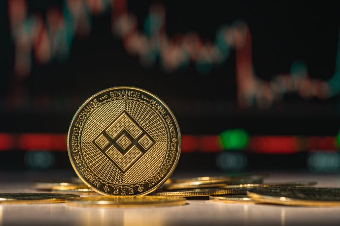 Eine goldene BNB-Kryptowährungsmünze steht aufrecht zwischen mehreren anderen Münzen, mit einem verschwommenen Aktiendiagramm und leuchtenden farbigen Lichtern im Hintergrund.