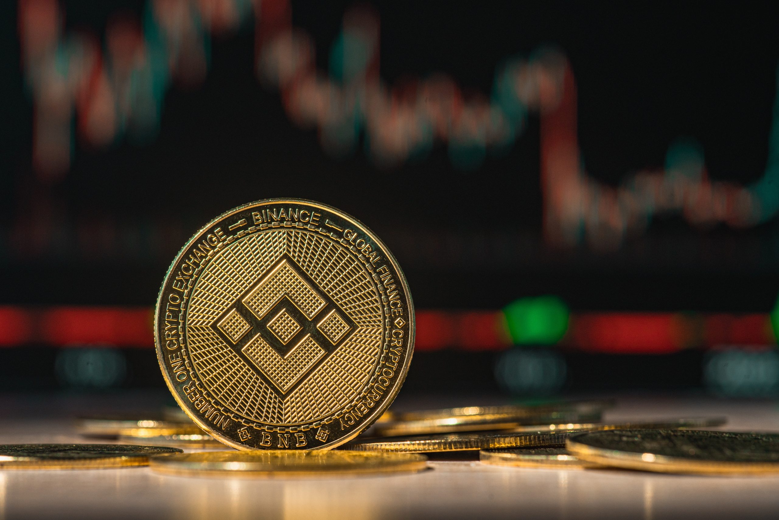 Eine goldene BNB-Kryptowährungsmünze steht aufrecht zwischen mehreren anderen Münzen, mit einem verschwommenen Aktiendiagramm und leuchtenden farbigen Lichtern im Hintergrund.