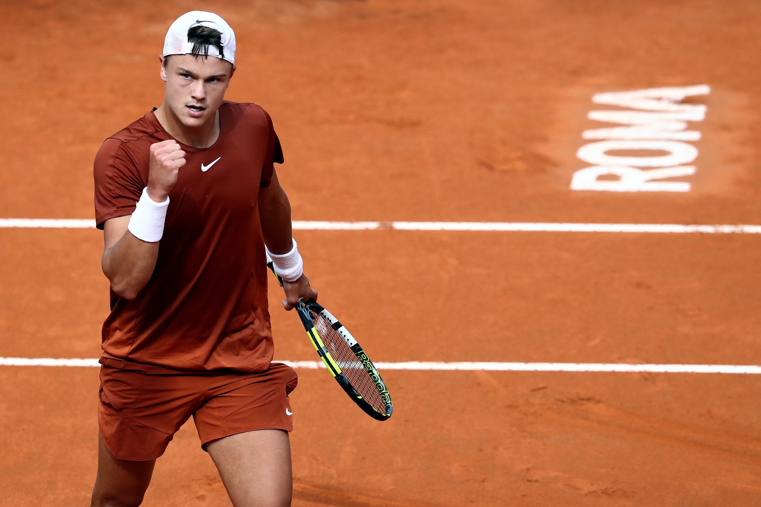 Ein Tennisspieler auf einem Sandplatz feiert triumphierend das Tennisturnier. Mit geballter Faust, in braunem Outfit und weißer Kappe, hält er einen Schläger in der Hand. ROMA ist auf dem Platz deutlich zu sehen.