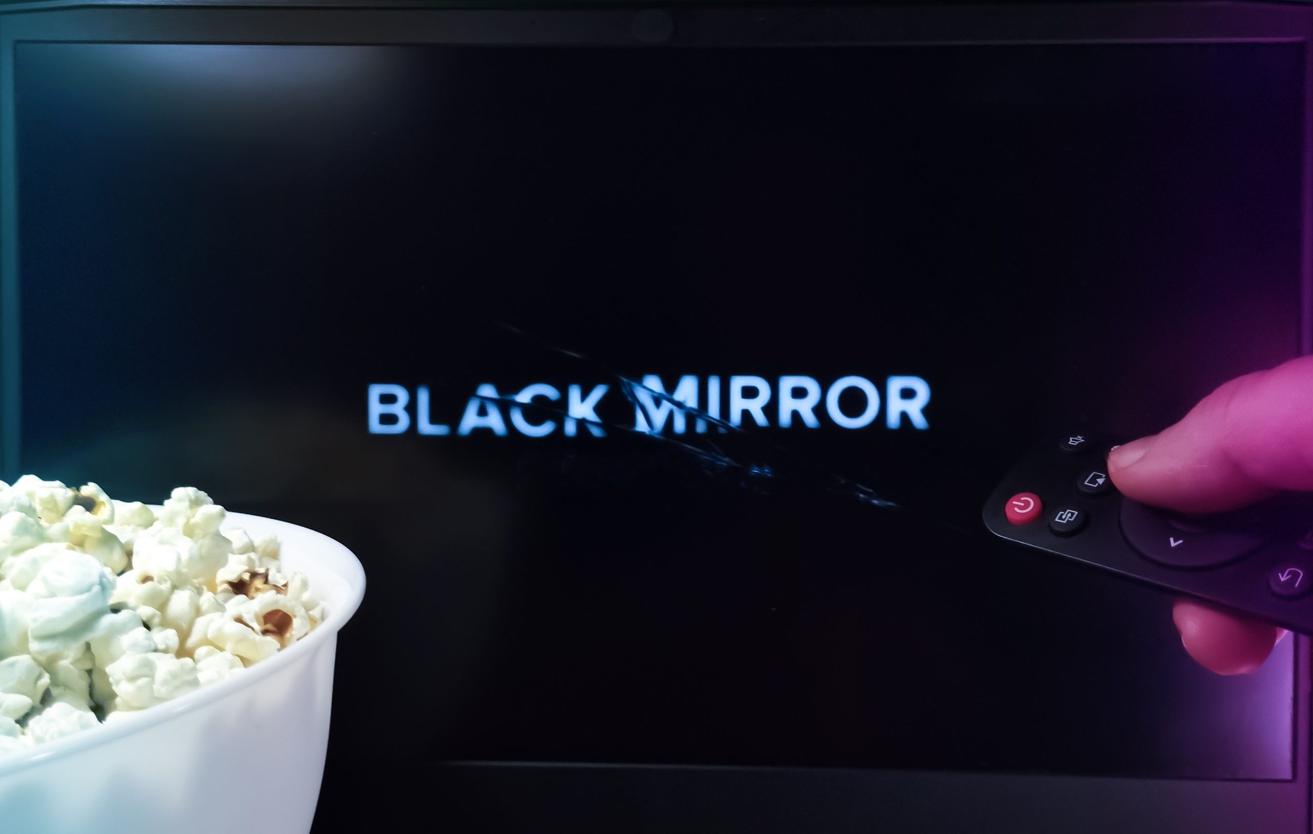 Eine Hand hält eine Fernbedienung vor einem Bildschirm, auf dem „Black Mirror“ läuft, am Rand des Bildschirms ist „Pump.fun“ zu sehen und daneben weckt eine Schüssel Popcorn Vorfreude.