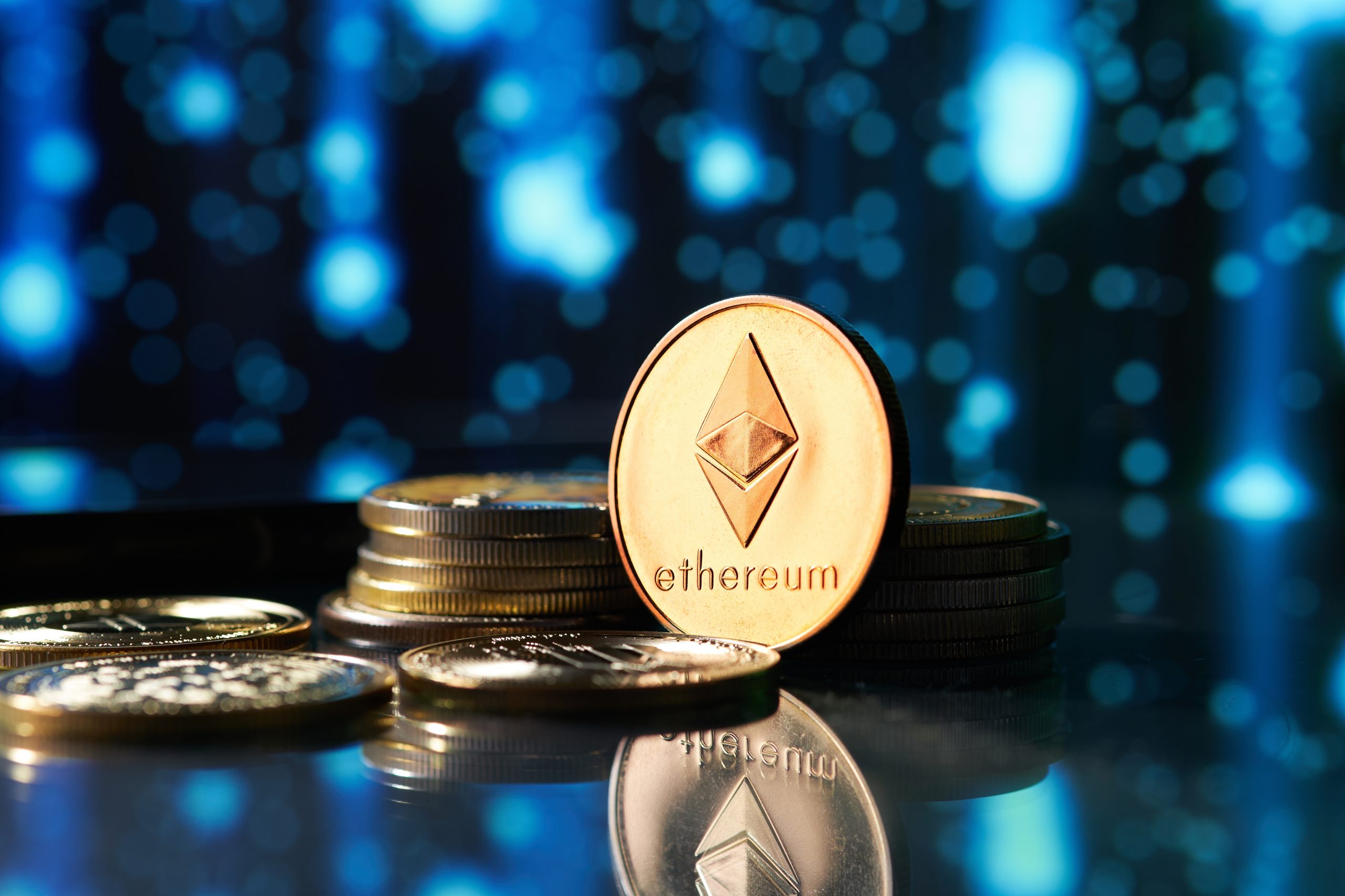Mehrere physische Ethereum-Münzen mit dem ikonischen Logo sind auf einer reflektierenden Oberfläche vor einem verschwommenen blau-schwarzen Hintergrund gestapelt und verstreut.