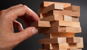 Eine Hand zieht vor einem schlichten, dunklen Hintergrund einen Holzblock aus einem gestapelten Turmspiel, ähnlich wie Jenga