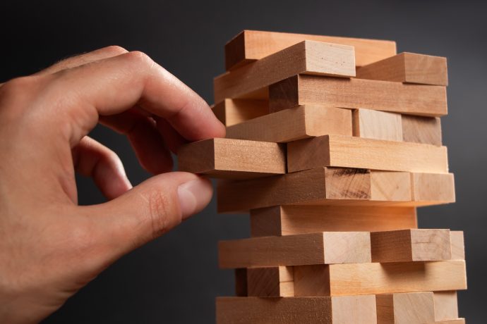 Eine Hand zieht vor einem schlichten, dunklen Hintergrund einen Holzblock aus einem gestapelten Turmspiel, ähnlich wie Jenga