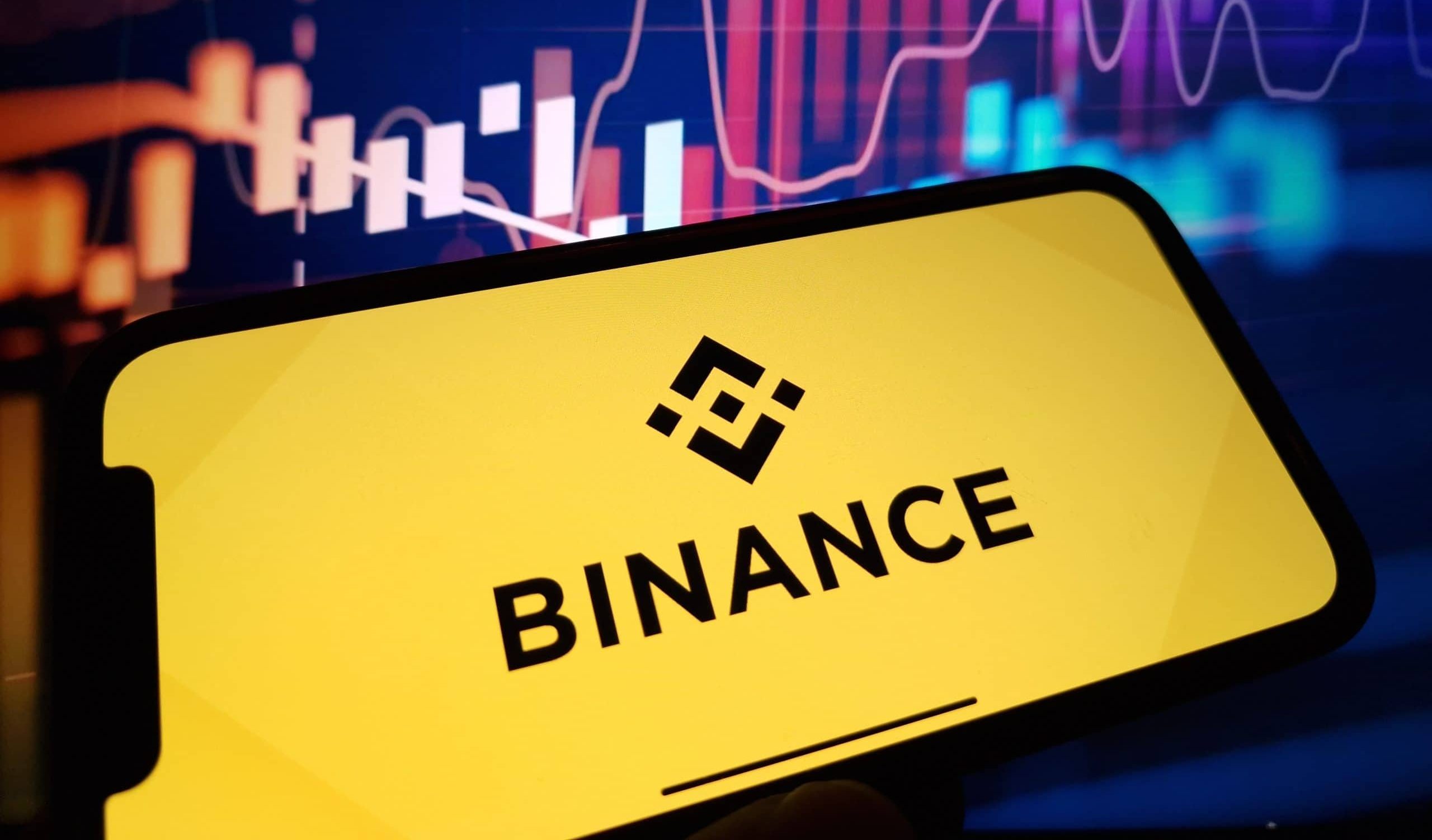 Ein Smartphone mit dem Binance-Logo steht prominent vor einem Bildschirm voller dynamischer Finanzdiagramme und Aktiencharts und verkörpert den Puls der digitalen Finanzwelt.