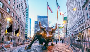 Eine große bronzene Bullenskulptur steht auf einer Kopfsteinpflasterstraße zwischen hohen Gebäuden, im Hintergrund sind zwei amerikanische Flaggen und ein dezentes Bitcoin-Symbol zu sehen.