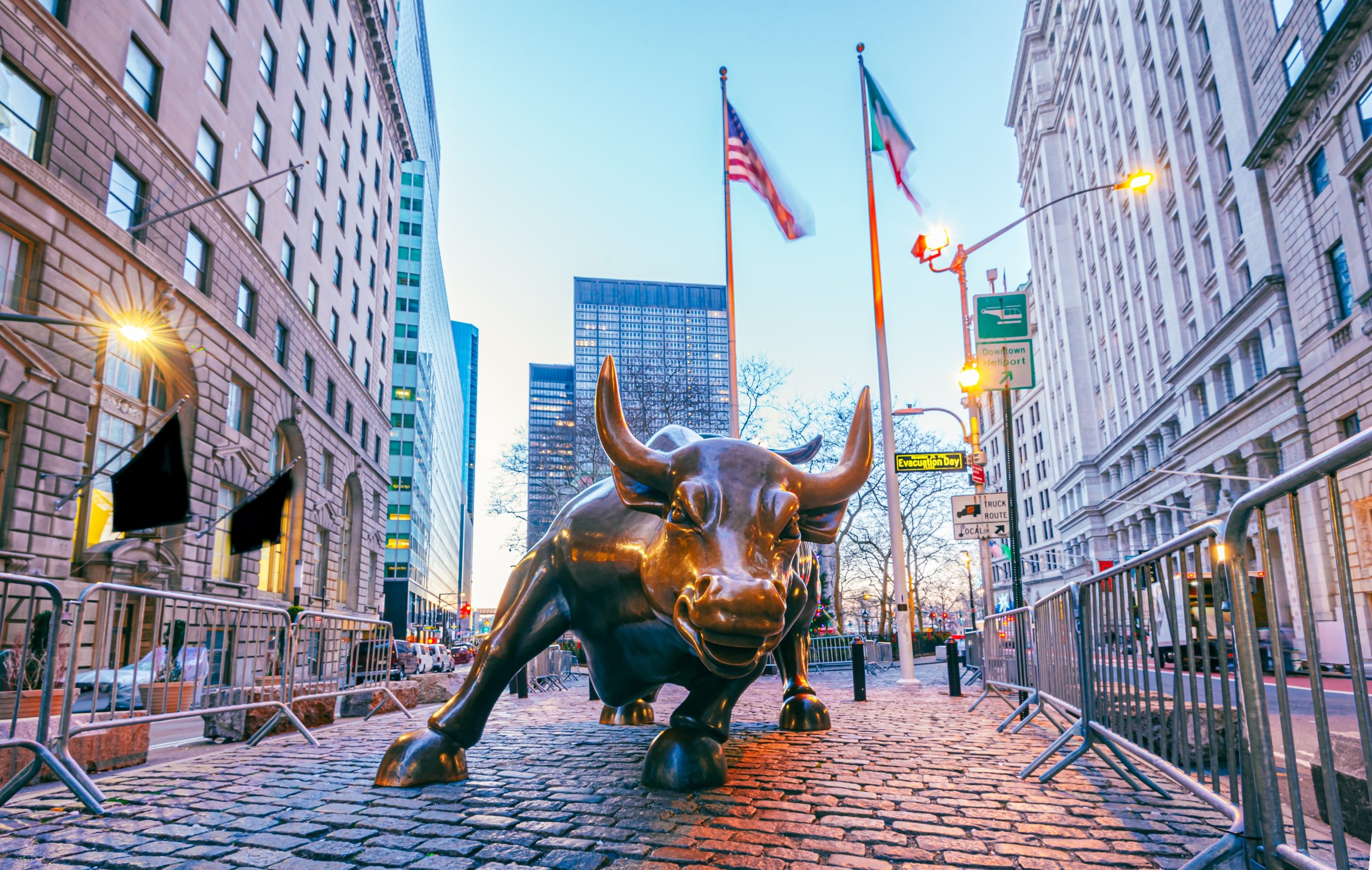 Eine große bronzene Bullenskulptur steht auf einer Kopfsteinpflasterstraße zwischen hohen Gebäuden, im Hintergrund sind zwei amerikanische Flaggen und ein dezentes Bitcoin-Symbol zu sehen.