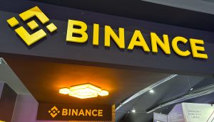 Ein Schild mit der Aufschrift „BINANCE“ in fetten gelben Buchstaben und einem darüber angebrachten Logo ist bei einer Veranstaltung prominent auf einer schwarzen Struktur angebracht. Durch beleuchtete Deckenleuchten hervorgehoben, unterstreicht das Schild dezent die Rolle von Binance bei Spot-Handel-Transaktionen.