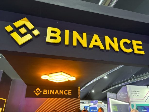 Ein Schild mit der Aufschrift „BINANCE“ in fetten gelben Buchstaben und einem darüber angebrachten Logo ist bei einer Veranstaltung prominent auf einer schwarzen Struktur angebracht. Durch beleuchtete Deckenleuchten hervorgehoben, unterstreicht das Schild dezent die Rolle von Binance bei Spot-Handel-Transaktionen.