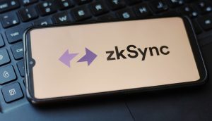 Ein Smartphone mit dem Logo und Namen von zkSync liegt auf einer schwarzen Laptoptastatur und deutet auf die Erwartung eines bevorstehenden zkSync-Airdrops hin.