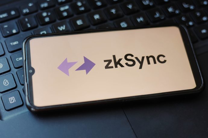 Ein Smartphone mit dem Logo und Namen von zkSync liegt auf einer schwarzen Laptoptastatur und deutet auf die Erwartung eines bevorstehenden zkSync-Airdrops hin.