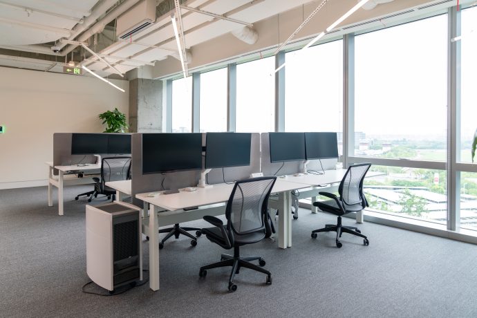 Ein leeres, modernes Büro, das für Krypto-Entwickler entworfen wurde, mit Schreibtischreihen mit ergonomischen Stühlen, Computern auf jedem Schreibtisch, großen Fenstern mit Blick auf die Stadt und einer freiliegenden Deckengestaltung.