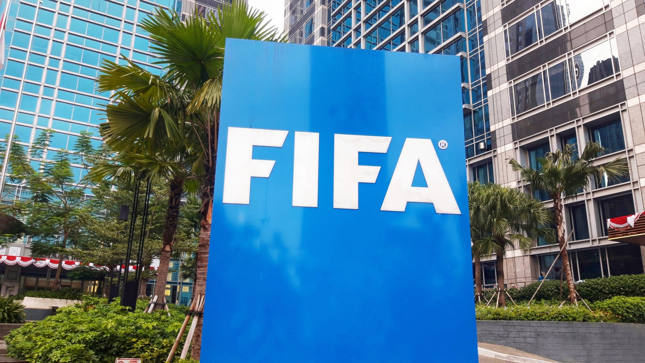 Vor modernen Bürogebäuden aus Glas und Palmen steht ein großes blaues Schild mit weißem FIFA-Schriftzug und deutet auf Innovationen hin, während die FIFA die Blockchain-Technologie erforscht.