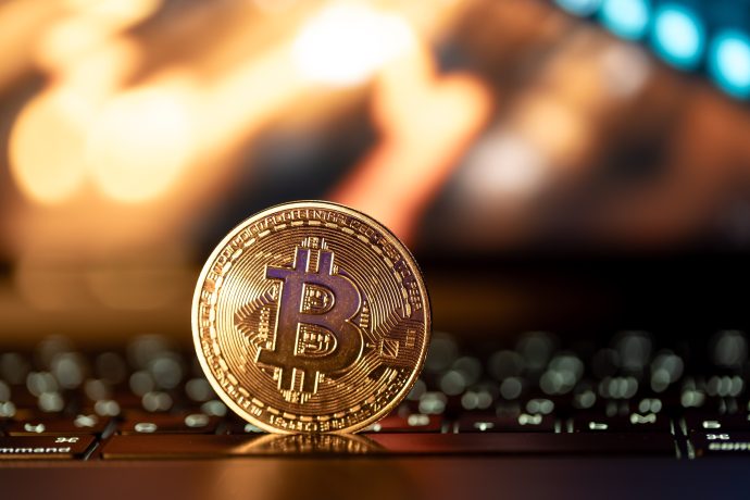 Eine physische Bitcoin-Münze steht aufrecht auf einer Laptop-Tastatur, mit verschwommenen, warm getönten Lichtern im Hintergrund, die den digitalen Aufstieg von Bitcoin symbolisieren.