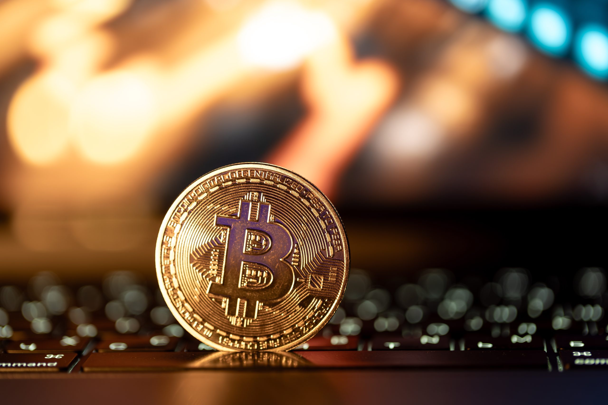 Eine physische Bitcoin-Münze steht aufrecht auf einer Laptop-Tastatur, mit verschwommenen, warm getönten Lichtern im Hintergrund, die den digitalen Aufstieg von Bitcoin symbolisieren.
