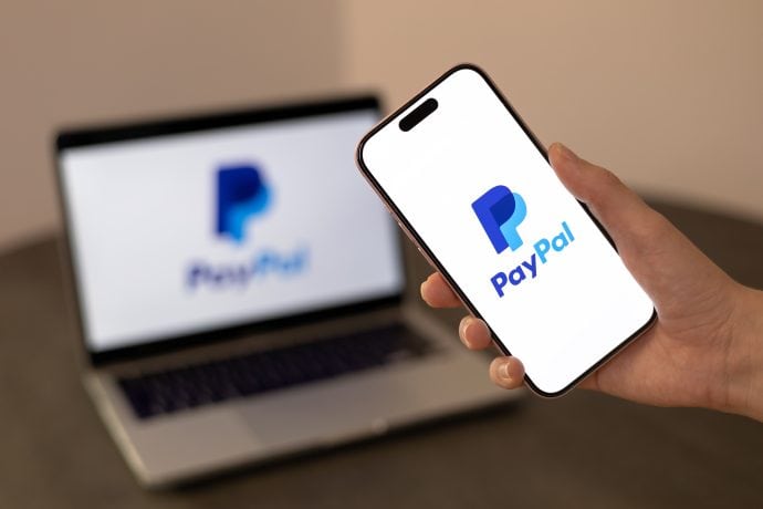 Eine Person hält ein Smartphone mit dem PayPal-Logo, während im Hintergrund auf einem Laptop-Bildschirm dasselbe Logo neben der Marke Solana angezeigt wird.