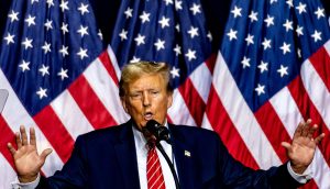 Donald Trump spricht mit erhobenen Händen in ein Mikrofon und diskutiert vor mehreren amerikanischen Flaggen über Bitcoin.