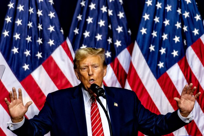 Donald Trump spricht mit erhobenen Händen in ein Mikrofon und diskutiert vor mehreren amerikanischen Flaggen über Bitcoin.