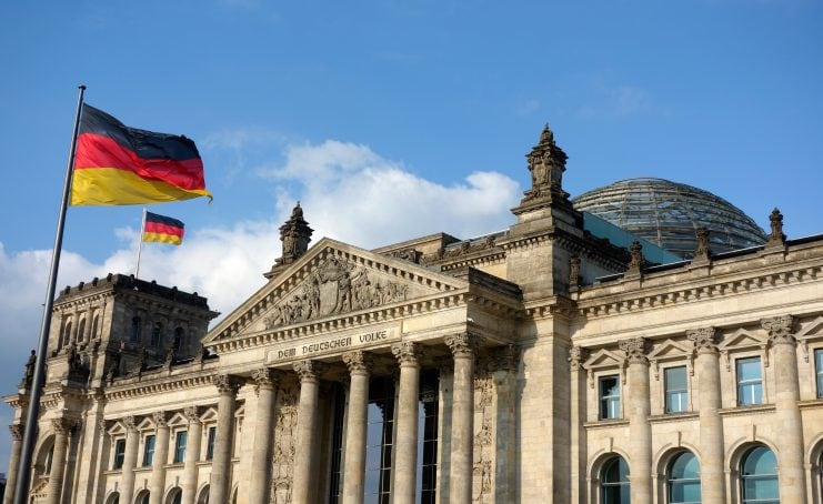 Das Reichstagsgebäude in Berlin thront stolz unter einem blauen Himmel mit vereinzelten Wolken. Zwei deutsche Flaggen – das Symbol der Einheit – wehen anmutig im Wind. Wie die Blockchain-Technologie steht das Gebäude für Transparenz und Sicherheit innerhalb seiner historischen Mauern.