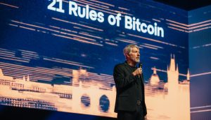 Ein Mann im schwarzen Anzug hält ein Mikrofon und steht auf der Bühne des BTC Prag vor einem großen Bildschirm, auf dem der Text „21 Regeln von Bitcoin“ angezeigt wird.