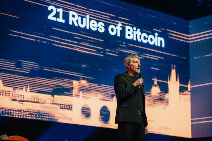 Ein Mann im schwarzen Anzug hält ein Mikrofon und steht auf der Bühne des BTC Prag vor einem großen Bildschirm, auf dem der Text „21 Regeln von Bitcoin“ angezeigt wird.