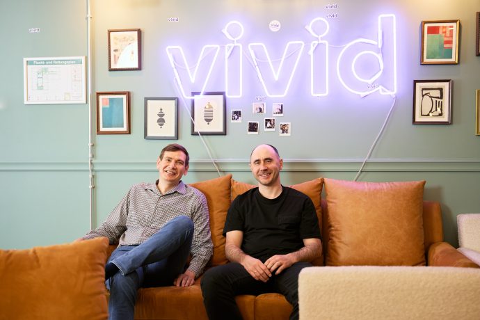 Zwei Männer sitzen auf einem hellbraunen Sofa vor gerahmter Kunst und einem großen, leuchtenden Neonschild und verleihen der modernen Lounge ein inspiriertes Aussehen – perfekt für ein Brainstorming oder die Durchsicht eines automatisch gespeicherten Entwurfs in entspannter Atmosphäre.