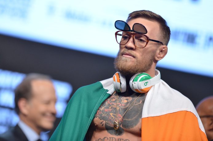 Ein Mann mit Bart trägt eine Doppelsonnenbrille, große Kopfhörer und eine irische Flagge über den Schultern – ganz im Stil von Conor McGregor –, während er bei einer Veranstaltung auf der Bühne steht.