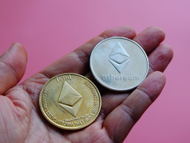 Eine Hand hält zwei Ethereum-Münzen, eine aus Gold und eine aus Silber, mit sichtbaren Ethereum-Logos und -Inschriften vor einem rosa Hintergrund.