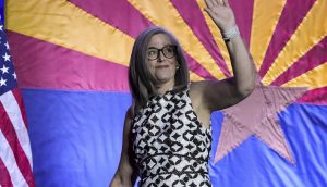 Eine Frau mit grauem Haar und Brille winkt, während sie vor einer Flagge des Staates Arizona und einer US-Flagge steht und bereit ist, ihre Anlagestrategie für die Navigation an der Börse in diesem Jahr zu besprechen.