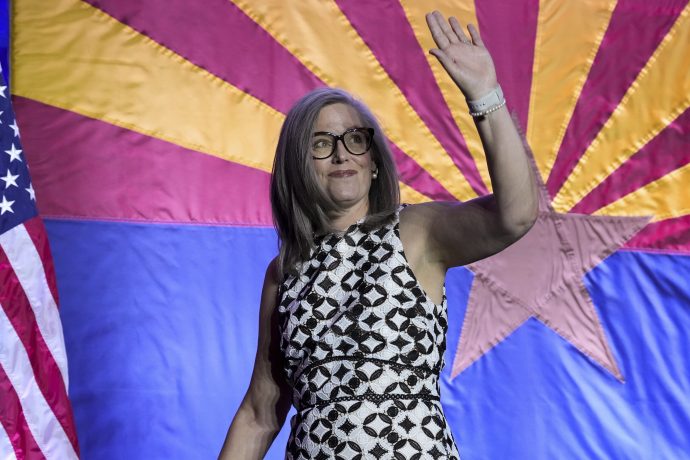 Eine Frau mit grauem Haar und Brille winkt, während sie vor einer Flagge des Staates Arizona und einer US-Flagge steht und bereit ist, ihre Anlagestrategie für die Navigation an der Börse in diesem Jahr zu besprechen.