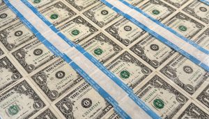 Blätter ungeschnittener Ein-Dollar-Scheine sind in Reihen angeordnet und durch blaue Klebestreifen voneinander getrennt, wodurch ein Kontrast zwischen traditioneller Währung und modernen digitalen Vermögenswerten wie Bitcoin entsteht.