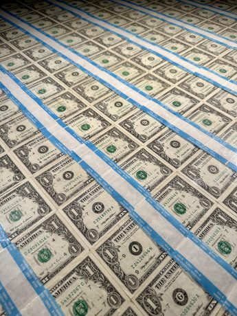 Blätter ungeschnittener Ein-Dollar-Scheine sind in Reihen angeordnet und durch blaue Klebestreifen voneinander getrennt, wodurch ein Kontrast zwischen traditioneller Währung und modernen digitalen Vermögenswerten wie Bitcoin entsteht.