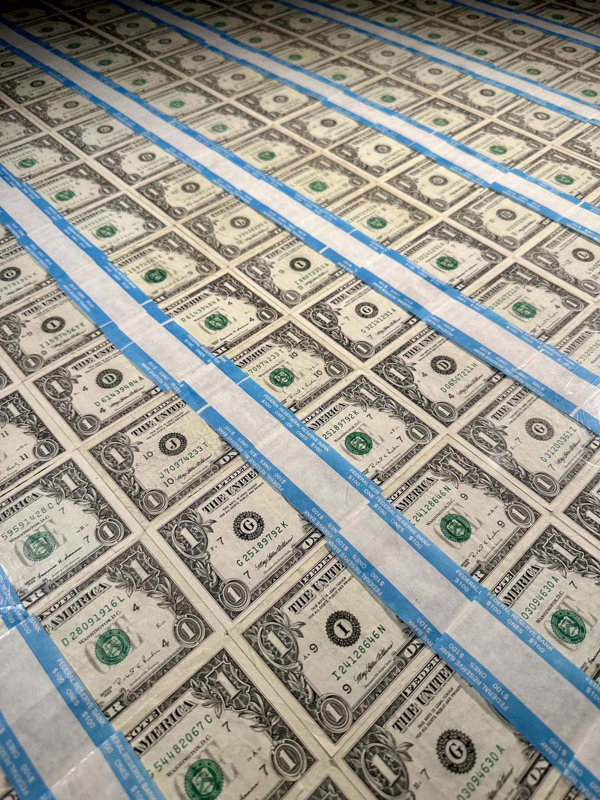 Blätter ungeschnittener Ein-Dollar-Scheine sind in Reihen angeordnet und durch blaue Klebestreifen voneinander getrennt, wodurch ein Kontrast zwischen traditioneller Währung und modernen digitalen Vermögenswerten wie Bitcoin entsteht.
