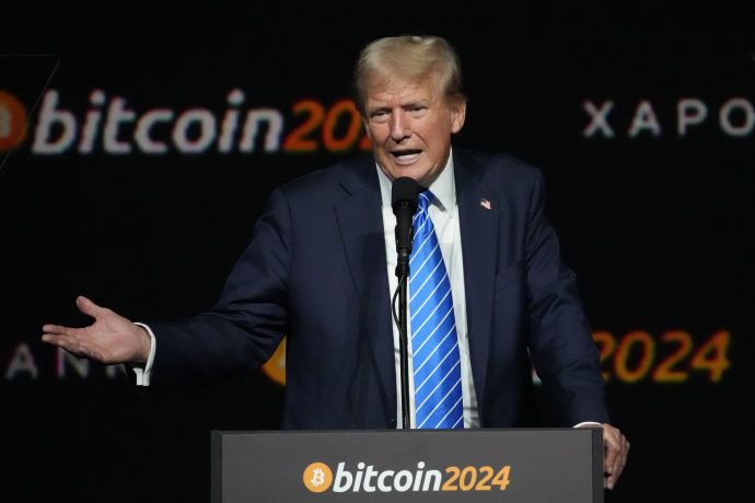 Ein Mann in Anzug und blau gestreifter Krawatte spricht an einem Podium mit der Aufschrift „Bitcoin 2024“ und einem schwarzen Hintergrund, auf dem „Bitcoin 2024“ und anderer Text zu sehen sind, während einer Veranstaltung, bei der Trumps Einfluss auf Kryptowährungen diskutiert wird.