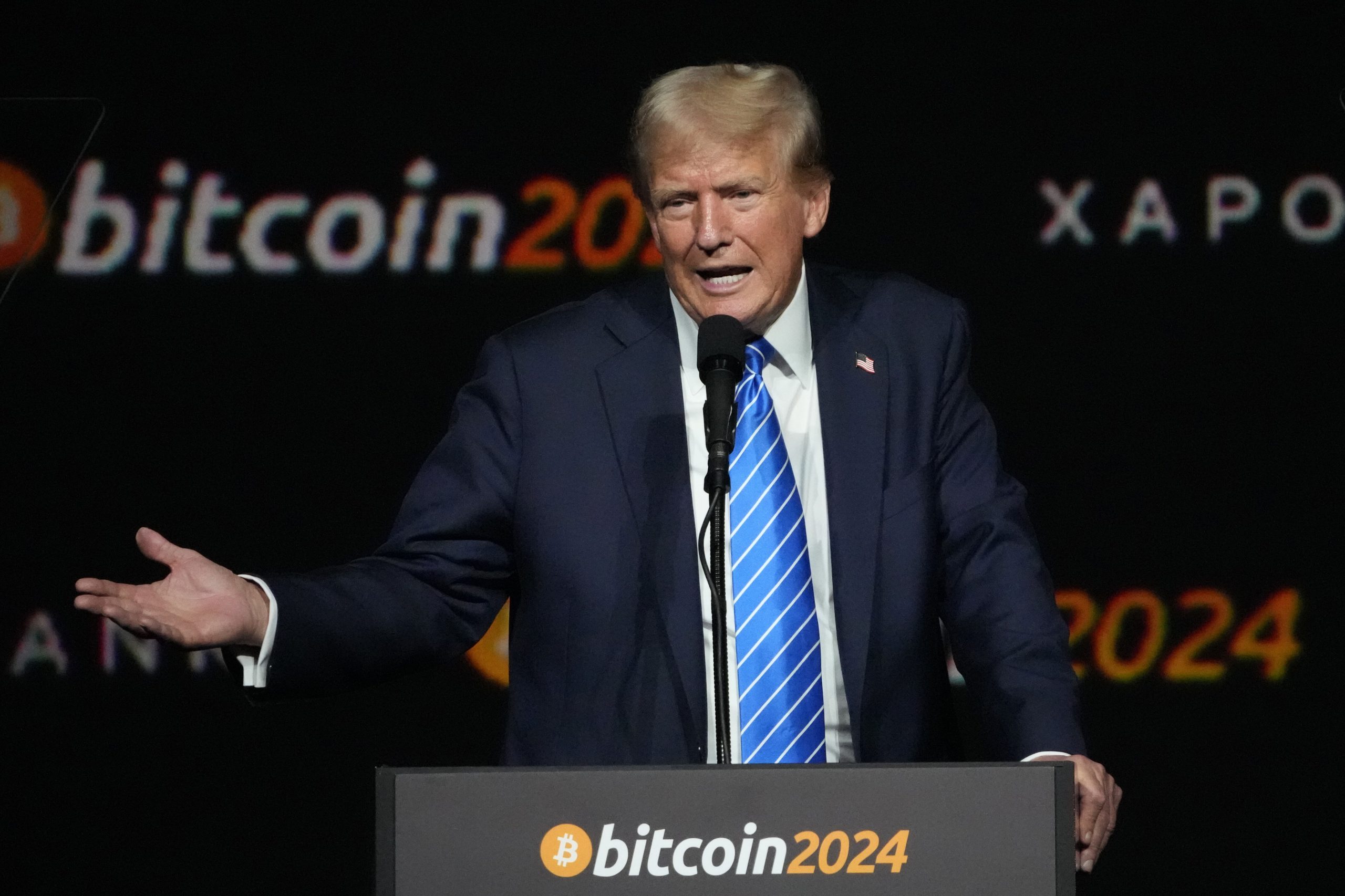Ein Mann in Anzug und blau gestreifter Krawatte spricht an einem Podium mit der Aufschrift „Bitcoin 2024“ und einem schwarzen Hintergrund, auf dem „Bitcoin 2024“ und anderer Text zu sehen sind, während einer Veranstaltung, bei der Trumps Einfluss auf Kryptowährungen diskutiert wird.