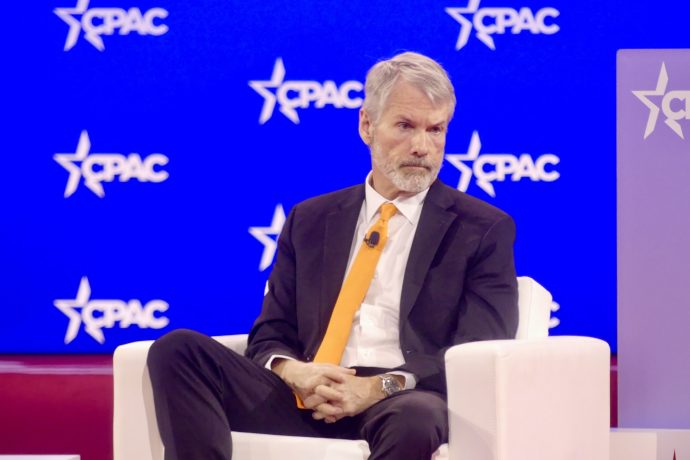 Ein Mann in Anzug und orangefarbener Krawatte sitzt auf einem weißen Stuhl vor einem blauen Hintergrund mit CPAC-Logo und diskutiert Themen wie Bitcoin und den Anstieg von Investitionen über 150.000 US-Dollar.