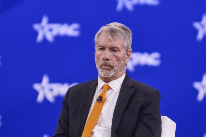 Ein Mann in Anzug und orangefarbener Krawatte sitzt auf der Bühne vor einem blauen Hintergrund mit PMC-Logos. Er hat graue Haare und einen Bart und trägt ein Mikrofon.