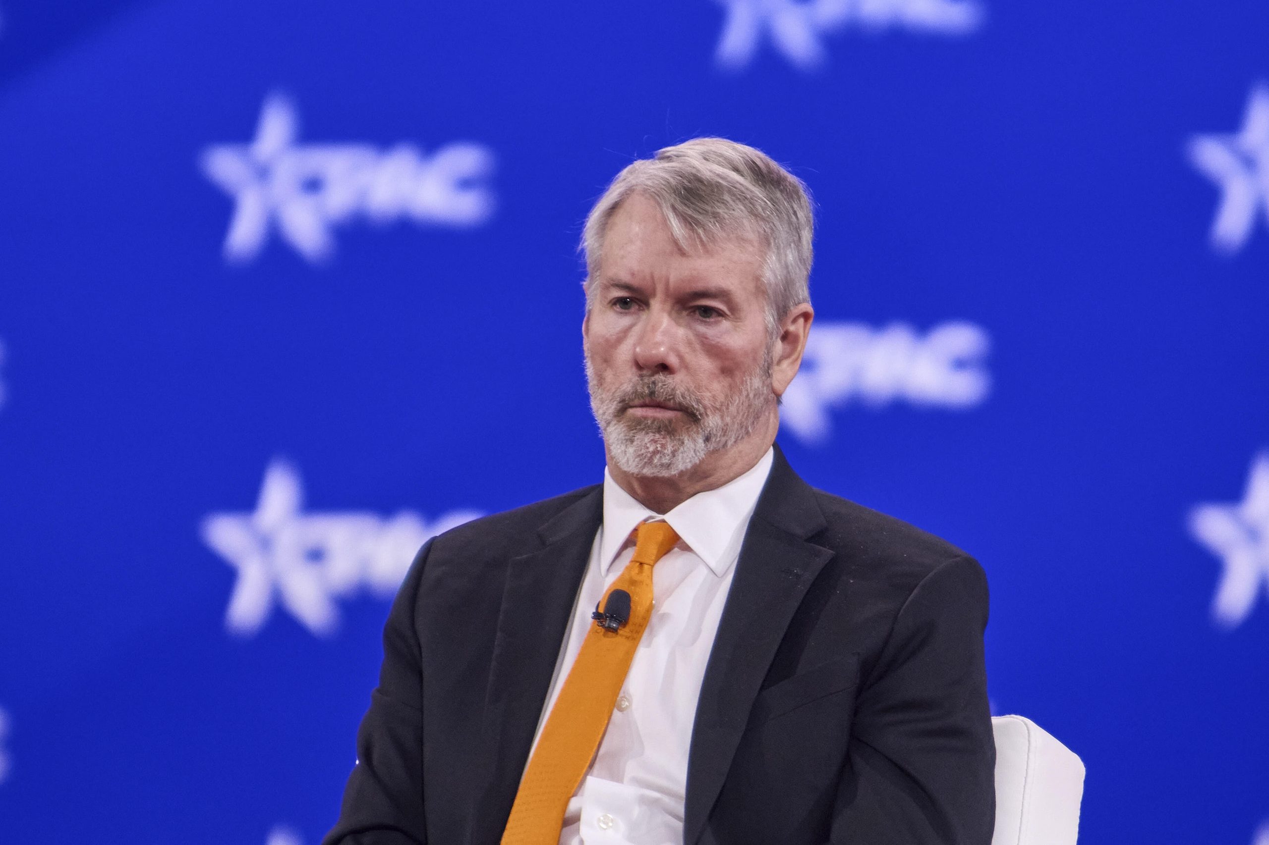 Ein Mann in Anzug und orangefarbener Krawatte sitzt auf der Bühne vor einem blauen Hintergrund mit PMC-Logos. Er hat graue Haare und einen Bart und trägt ein Mikrofon.
