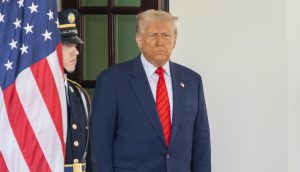 Ein Mann in dunklem Anzug und roter Krawatte, der Donald Trump ähnelt, steht neben einer amerikanischen Flagge und einem uniformierten Wachmann vor dem Eingang eines Gebäudes und erinnert damit an die Förmlichkeit einer Senatsveranstaltung.