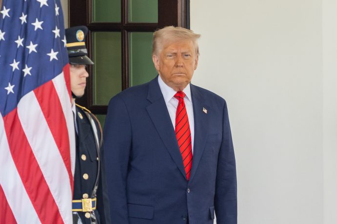 Ein Mann in dunklem Anzug und roter Krawatte, der Donald Trump ähnelt, steht neben einer amerikanischen Flagge und einem uniformierten Wachmann vor dem Eingang eines Gebäudes und erinnert damit an die Förmlichkeit einer Senatsveranstaltung.