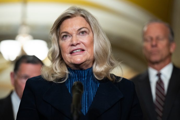 Senatorin Cynthia Lummis, mit langen blonden Haaren, spricht in ein Mikrofon, während im unscharfen Hintergrund im Innenbereich zwei Männer stehen.