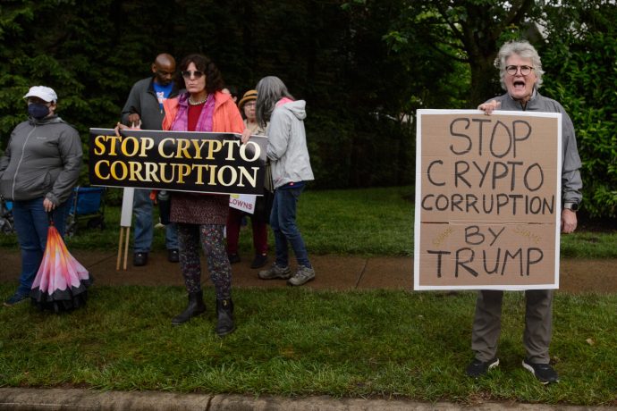 Menschen stehen im Freien und halten Schilder mit der Aufschrift „STOP KRYPT O CORRUPTION“ und „STOP CRYPTO CORRUPTION BY TRUMPS“.