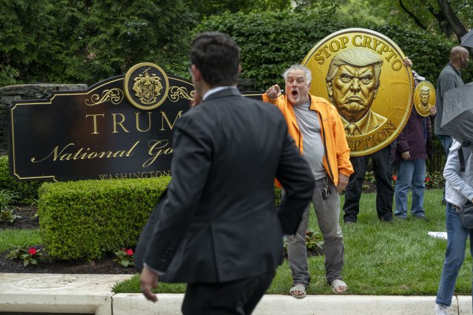 Ein Mann hält vor dem Trump National Golf Club ein großes, münzförmiges Schild mit Donald Trumps Gesicht und der Aufschrift „Stop Crypto Corruption“ hoch, während Passanten vorbeigehen.