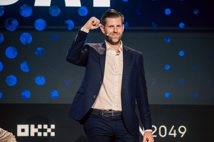 Eric Trump steht in einem marineblauen Anzug und einem hellen Hemd mit einem zur Faust erhobenen Arm auf der Bühne vor einem Hintergrund mit blauen Punkten und Veranstaltungslogos zu den Themen Banken und Kryptowährungen.