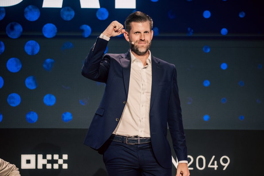 Eric Trump steht in einem marineblauen Anzug und einem hellen Hemd mit einem zur Faust erhobenen Arm auf der Bühne vor einem Hintergrund mit blauen Punkten und Veranstaltungslogos zu den Themen Banken und Kryptowährungen.