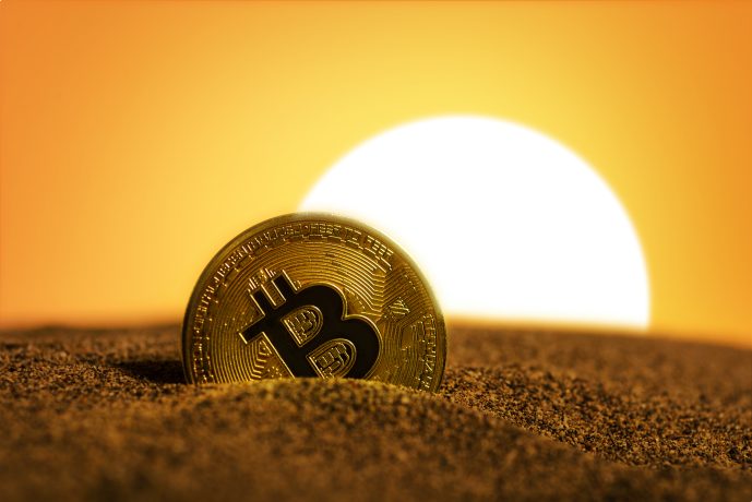 Eine glänzende, teilweise im Sand vergrabene Bitcoin-Münze schimmert unter der hellen Sonne, die im Hintergrund entweder untergeht oder aufgeht.