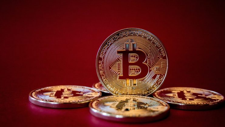 Fünf physische Bitcoin-Münzen werden vor einem schlichten roten Hintergrund angezeigt, wobei eine Bitcoin-Münze aufrecht in der Mitte steht.