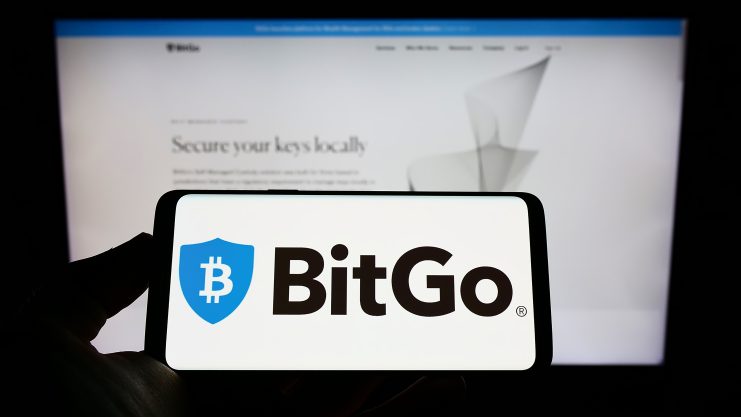 Auf einem Smartphone wird das BitGo-Logo vor einem unscharfen Computerbildschirm angezeigt, auf dem die Website von BitGo zu sehen ist, was die Genehmigung der MiCA-Lizenz durch die BaFin hervorhebt.